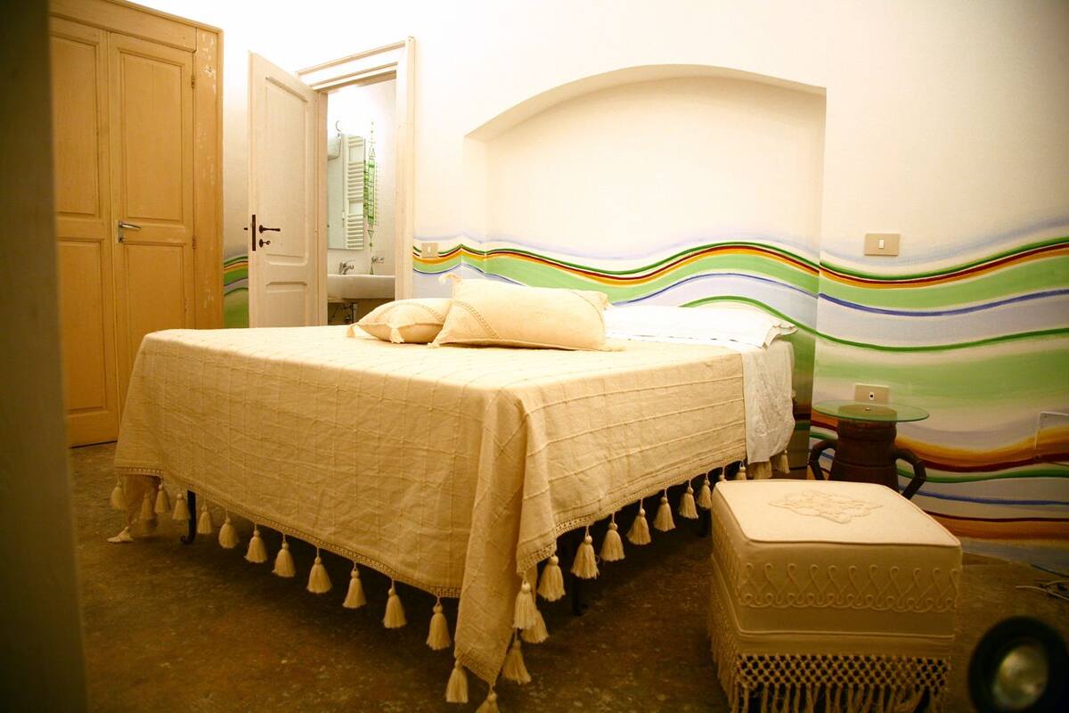 A bedroom with a bed, a nightstand, a chair, and a wall with a colorful design.