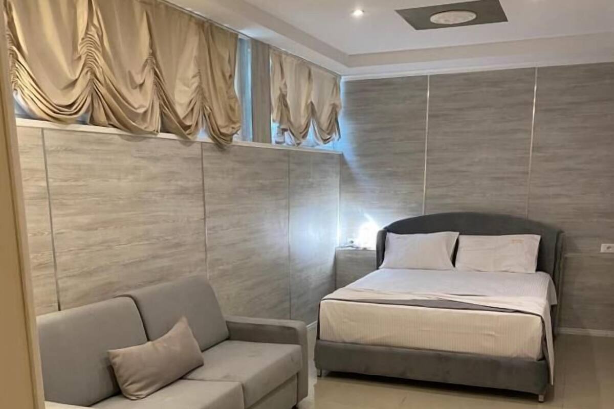 Un dormitorio moderno con un sofá gris, una cama con ropa de cama blanca y un ventanal con cortinas.