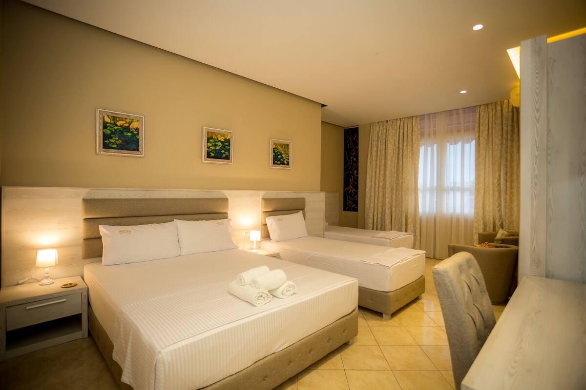 Una habitación de hotel con dos camas, una mesita de noche con lámpara, una silla y una obra de arte en la pared.