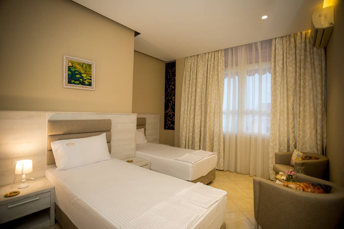 Habitación de hotel con dos camas, una mesita de noche con lámpara, una silla y un ventanal con cortinas.