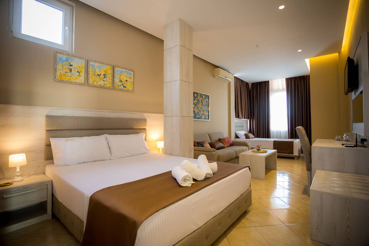 Habitación de hotel con una cama grande, un sofá, un escritorio y un televisor. Hay tres cuadros en la pared y una ventana que proporciona luz natural.