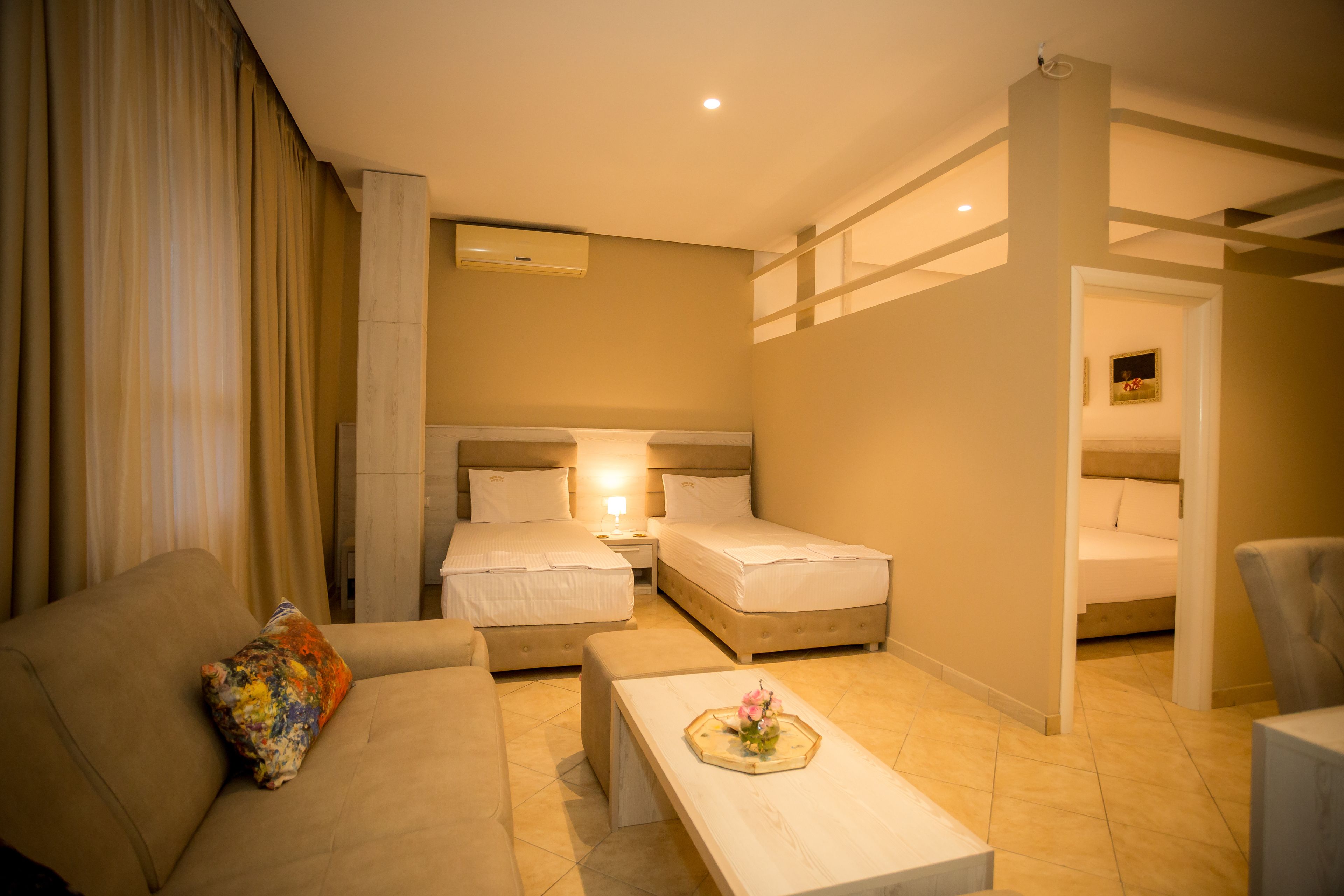 A hotel room with two beds, a sofa, a coffee table with a flower arrangement, and a nightstand with a lamp.