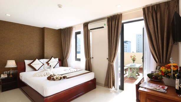 A hotel room with a large bed, a nightstand, a table with a bottle and glasses, and a view of the city through the window. - Kingdom Hotel (Da Nang)
