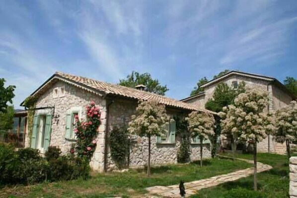 A stone house with a tiled roof, green shutters, and a garden with flowering plants.