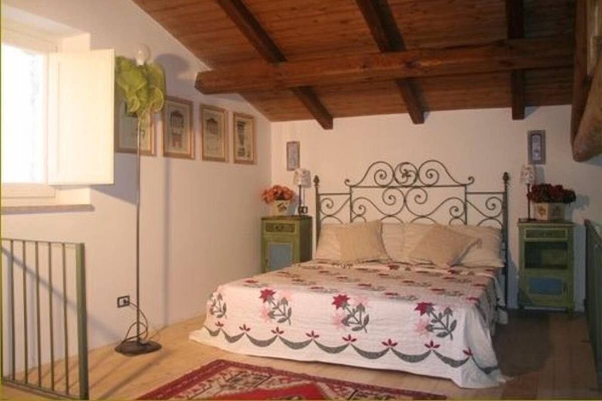 Une chambre avec un plafond en bois, un lit recouvert d’une literie à motifs floraux, deux tables de chevet avec des lampes et une fenêtre avec un rideau.