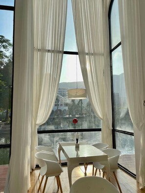 A room with a large window, white curtains, a table with chairs, and a view of a body of water and buildings.