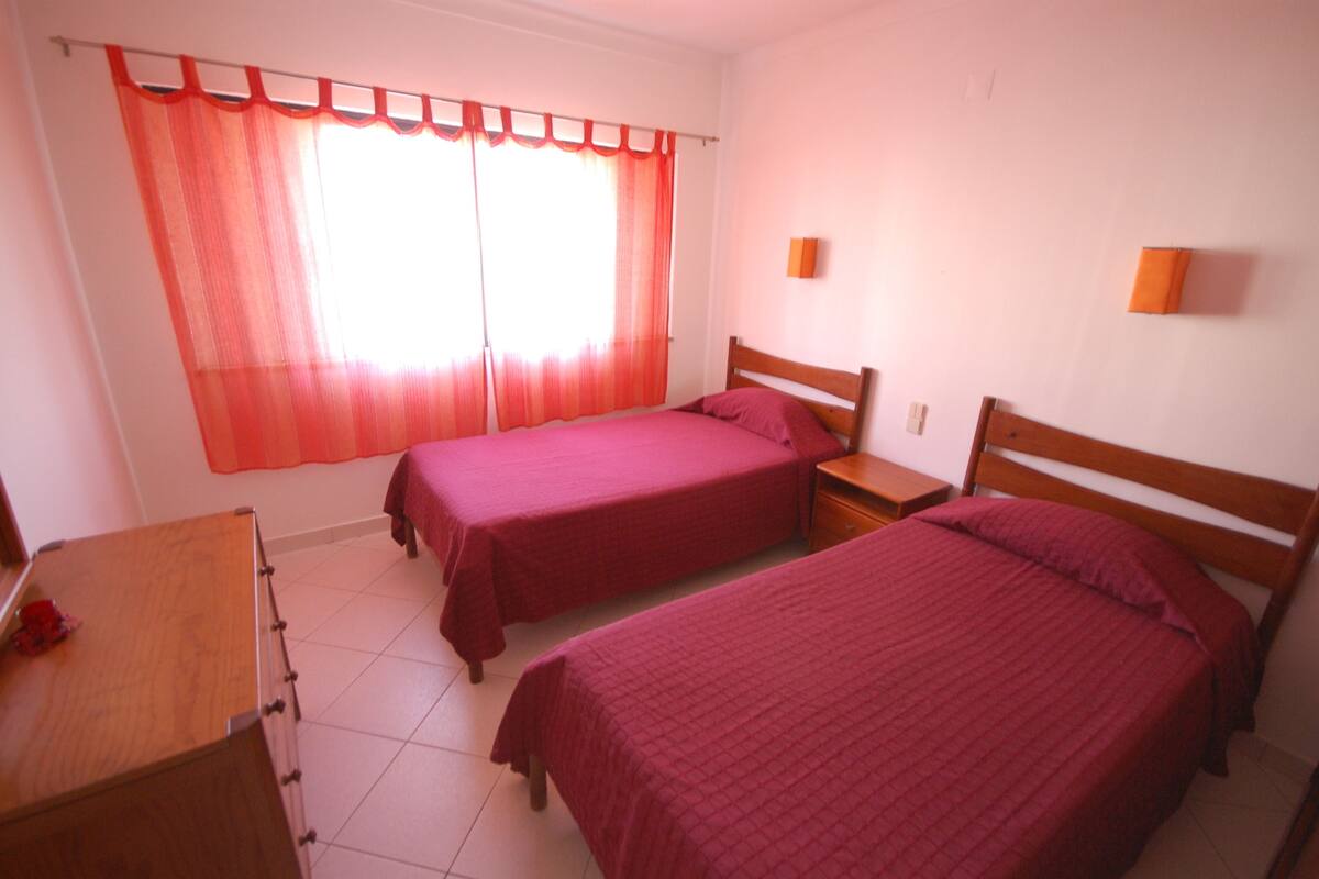 A room with two single beds, a wooden desk, a small table, and a window with red curtains.
