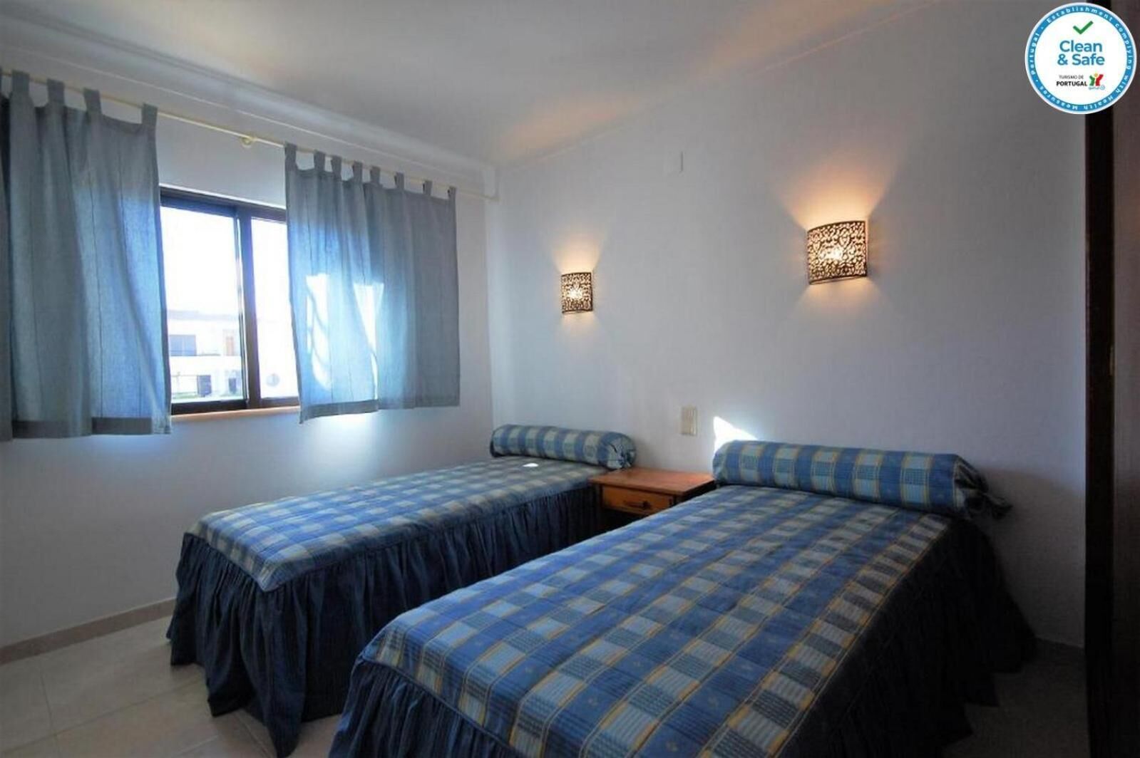 A room with two beds, a window with curtains, and wall-mounted lamps.