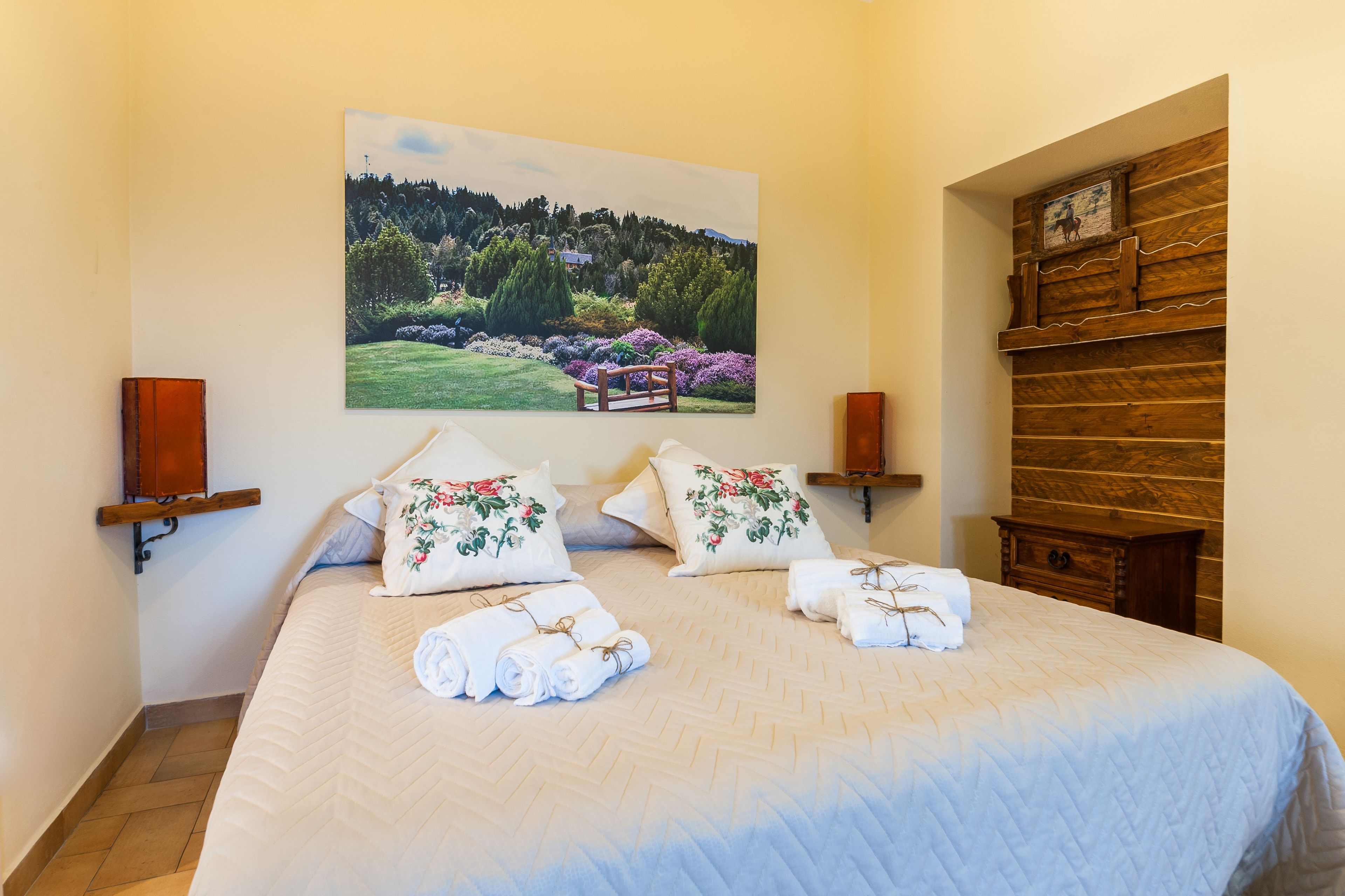 Una cama bien hecha con almohadas y toallas florales, un cuadro de un paisaje en la pared y un estante de madera con una fotografía.