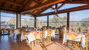 Un área de comedor al aire libre con mesas y sillas de madera, ventanas amplias y vistas del paisaje circundante.