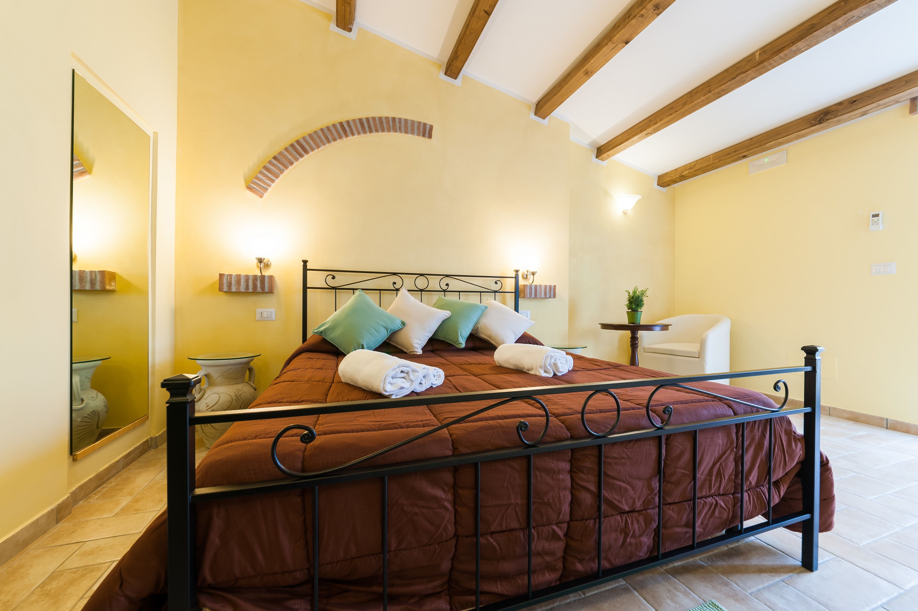 A bed with a metal frame, a headboard, and a brown quilt. There is a bedside table with a lamp and a small potted plant. The room has a tiled floor and a yellow wall with a wooden ceiling beam.