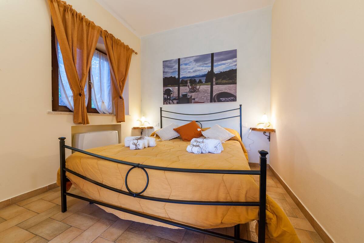 Una cama bien hecha con cabecera, una ventana con cortinas, un cuadro colgado en la pared y lámparas de noche.