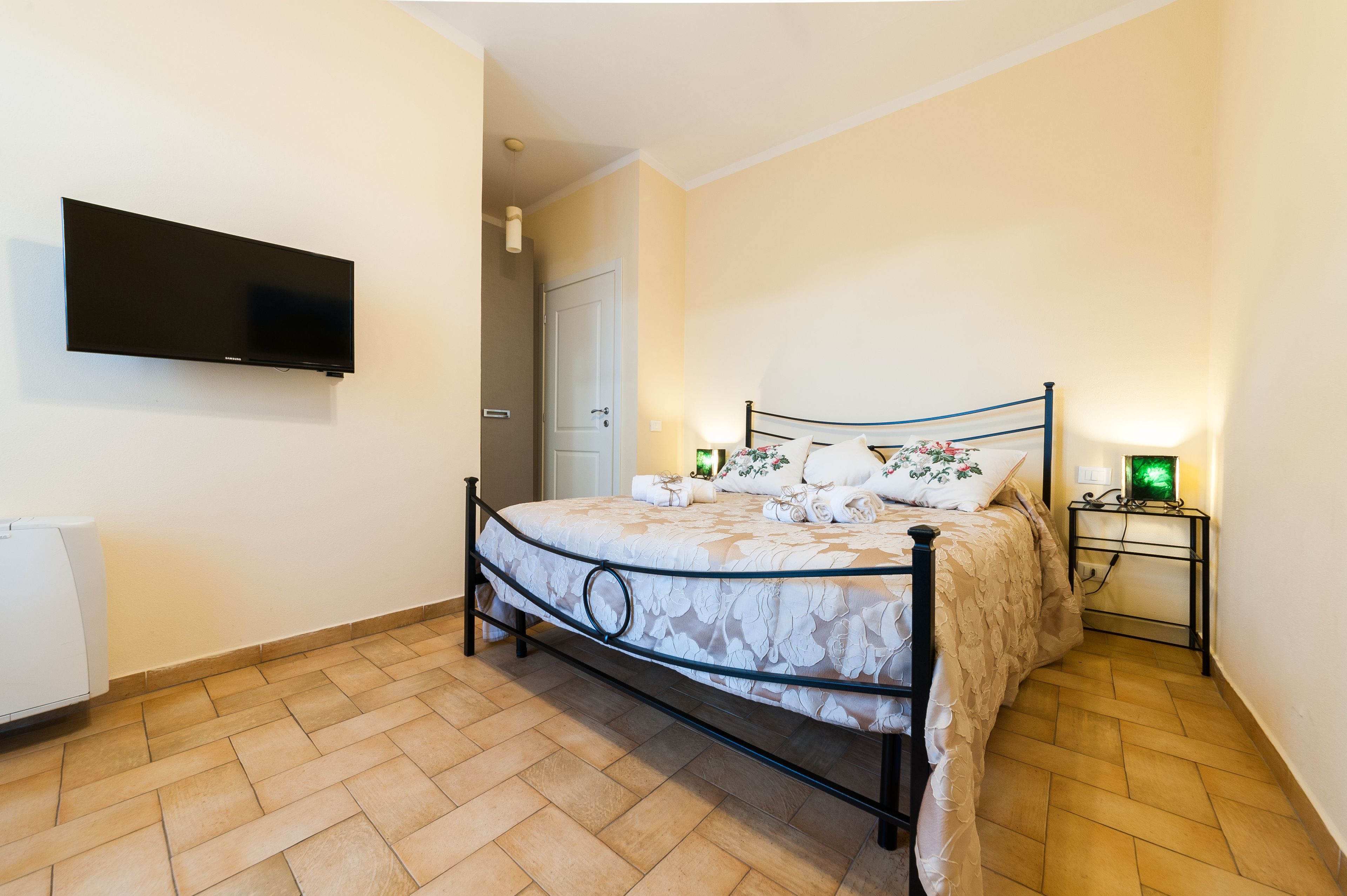 Un dormitorio con cama, televisor de pantalla plana montado en la pared, mesita de noche con lámpara y una puerta que da a otra habitación.