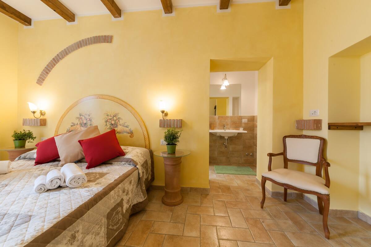 Un dormitorio con cama, almohadas, lámparas de noche, una silla, mesita de noche y baño con lavabo y espejo.