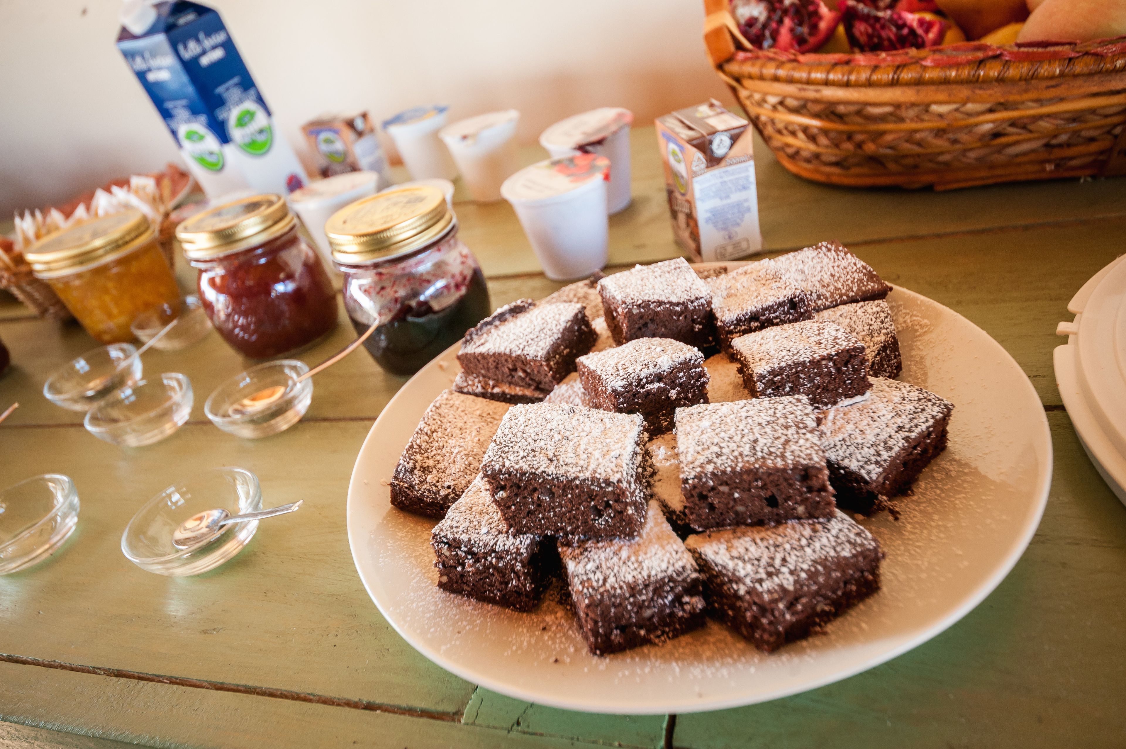 Un plato de brownies sobre una mesa con frascos de mermelada y una cesta de frutas al fondo.
