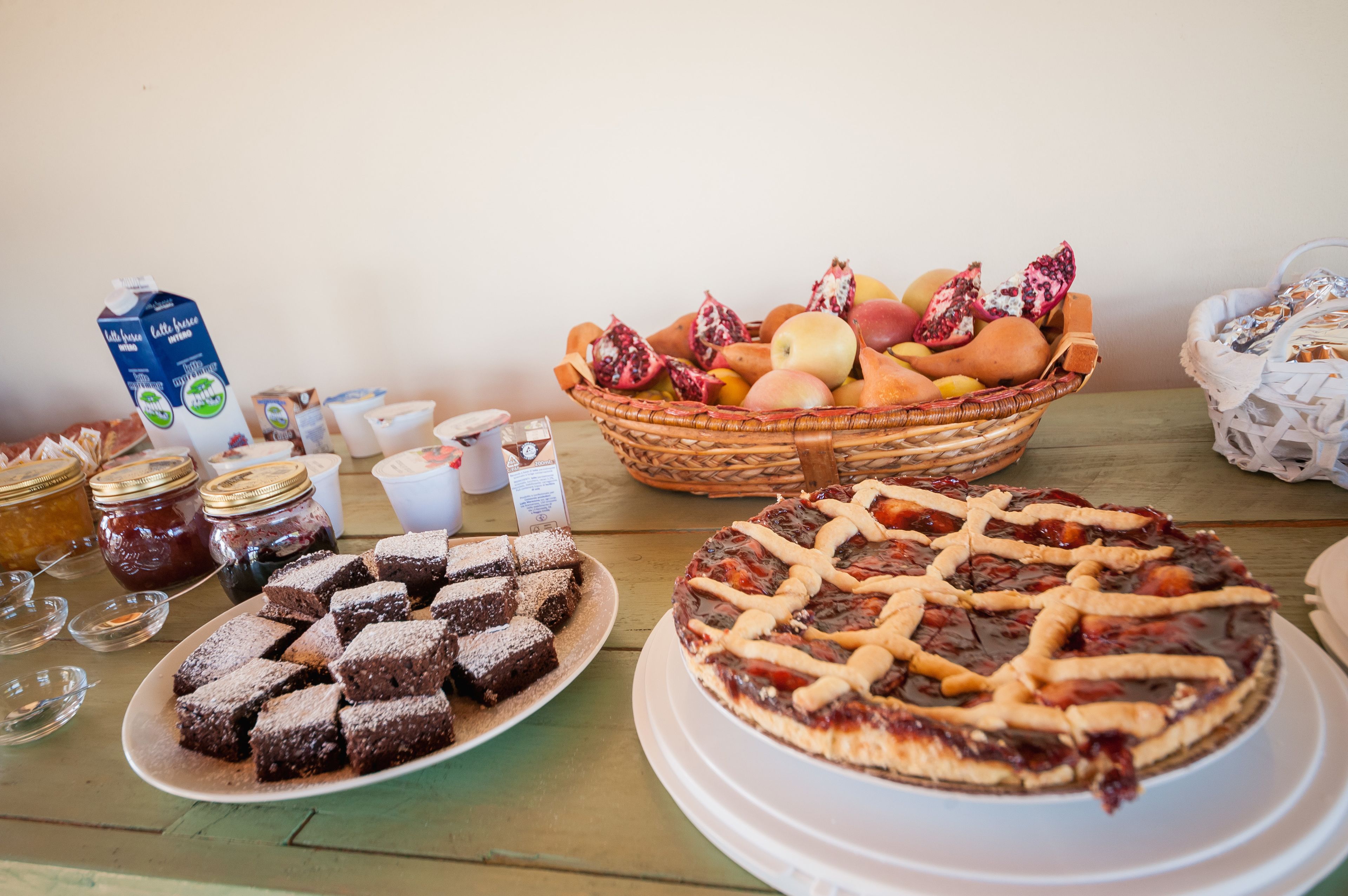 A table with a pie, a basket of fruits, jars of preserves, and a carton of milk.
