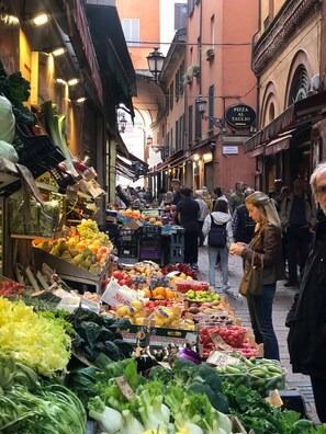 A bustling outdoor market with various stalls selling fresh produce and a sign for a pizza place.