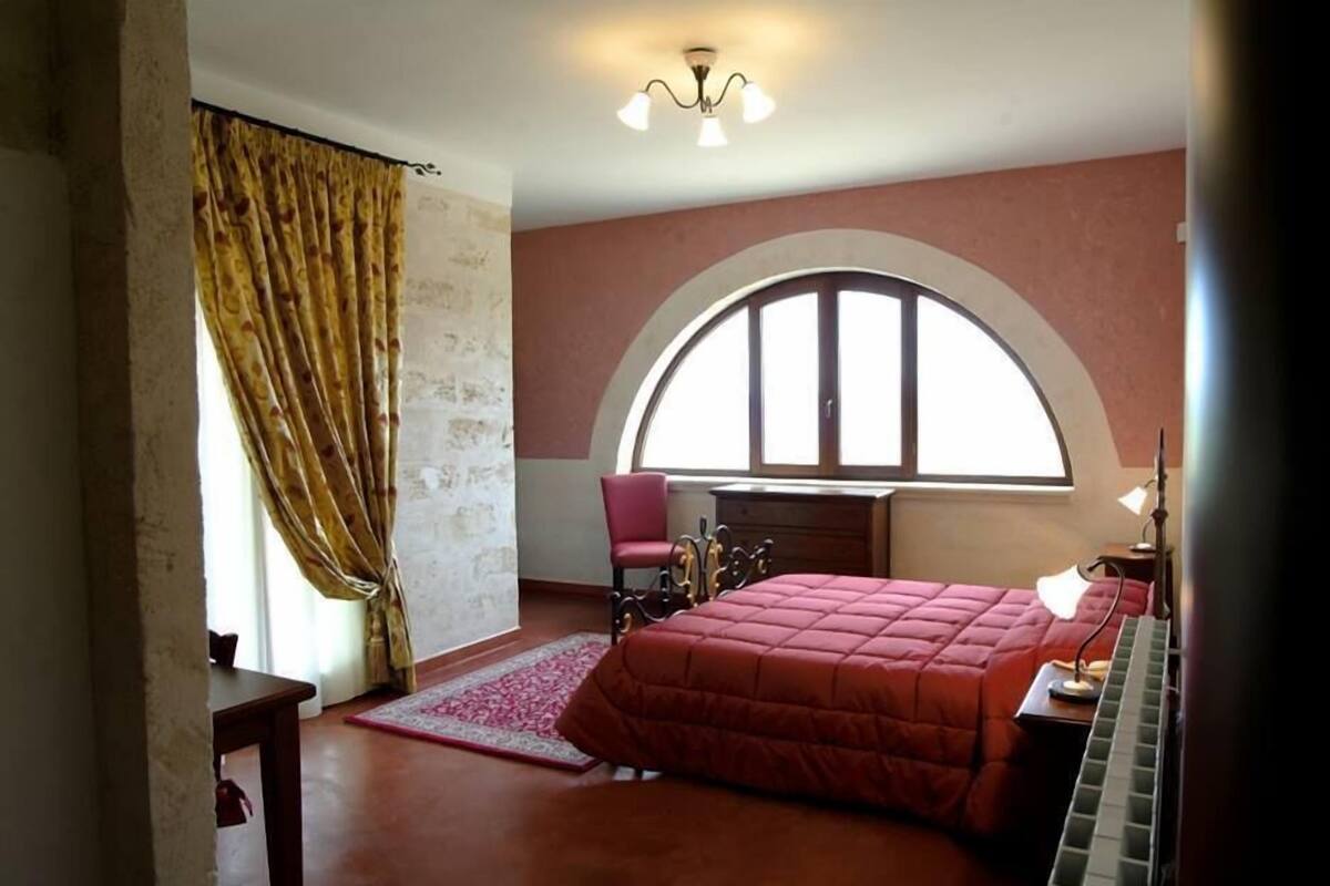 Une chambre à coucher avec un grand lit, un bureau, une chaise, une fenêtre en arc de cercle et un lustre.
