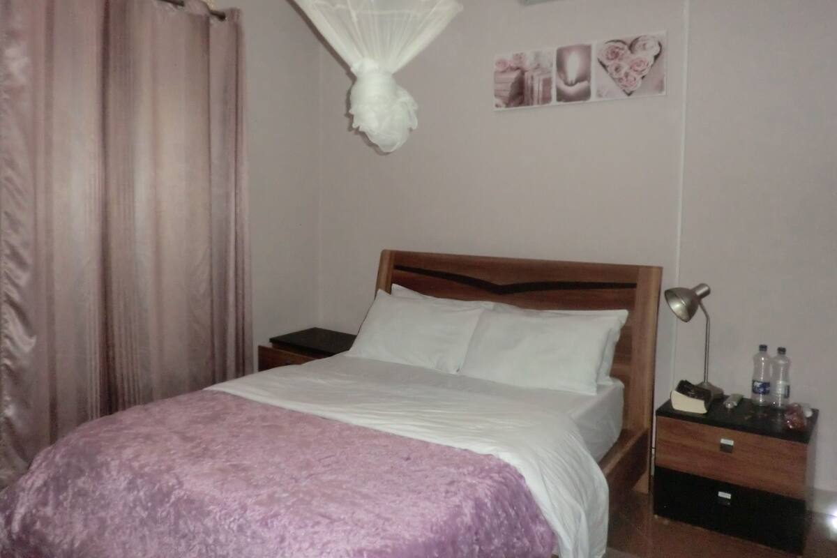 A bedroom with a bed, a bedside table, a nightstand, a lamp, and a wall-mounted air conditioner.