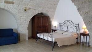 A bedroom with a stone wall, a bed with a metal frame, a wooden wardrobe, a blue sofa, and a bedside table with a lamp. - Masseria Ciavea (Gioia del Colle)