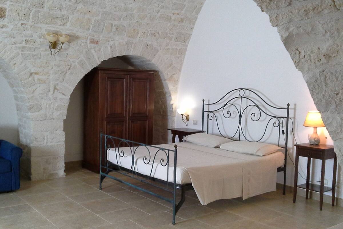 Ein Schlafzimmer mit Steingemäuer, einem Bett mit Metallgestell, einem Holzschrank, einem blauen Sofa und einem Nachttisch mit Lampe.