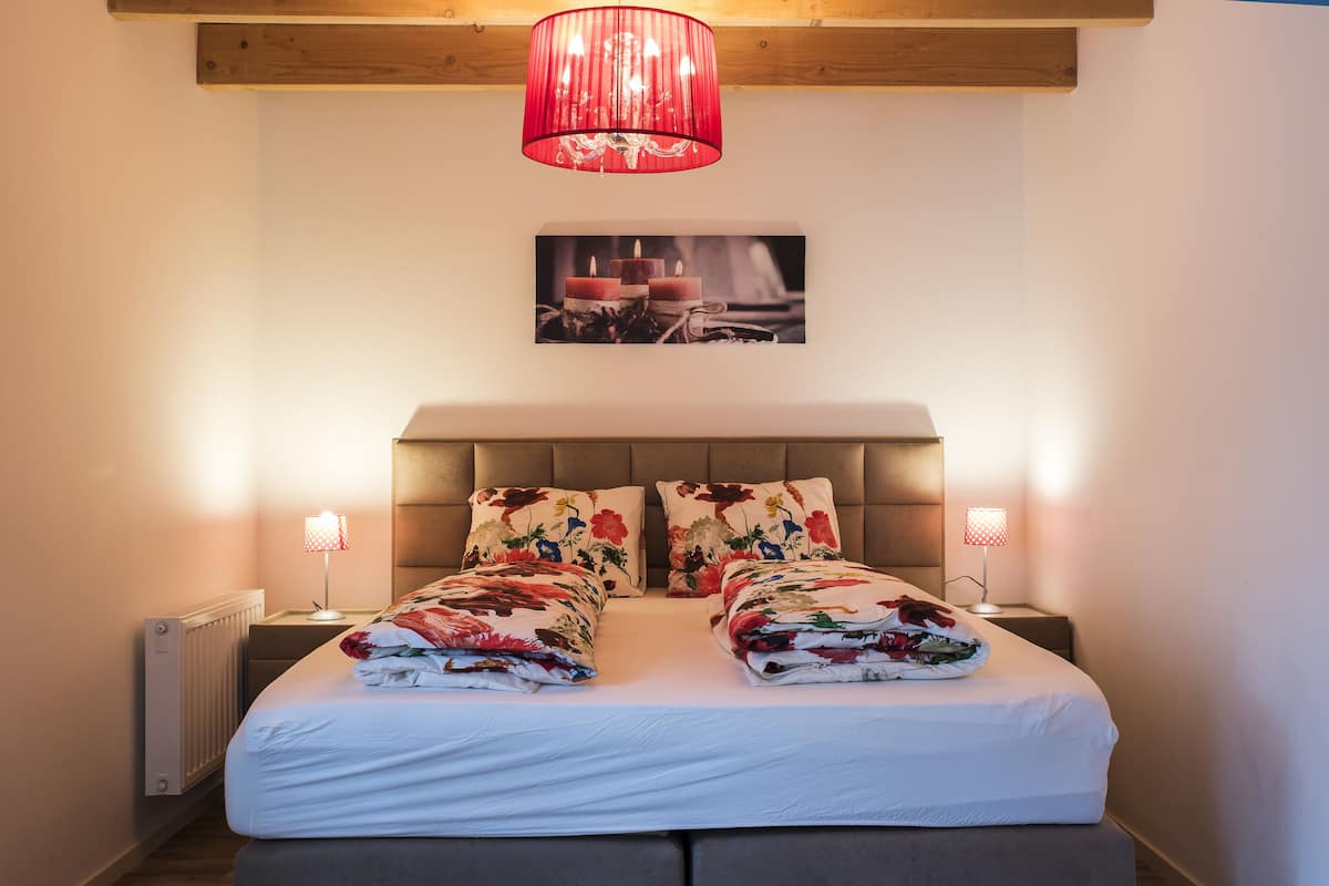 A neatly made bed with floral bedding, two bedside lamps, a red pendant light, and a framed picture on the wall.