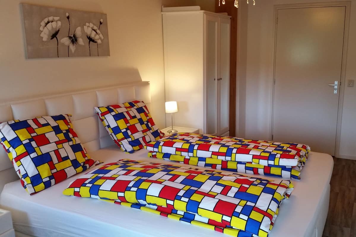 A bedroom with two beds, each with colorful geometric-patterned bedding. There is a nightstand with a lamp, a painting on the wall, and a wardrobe.
