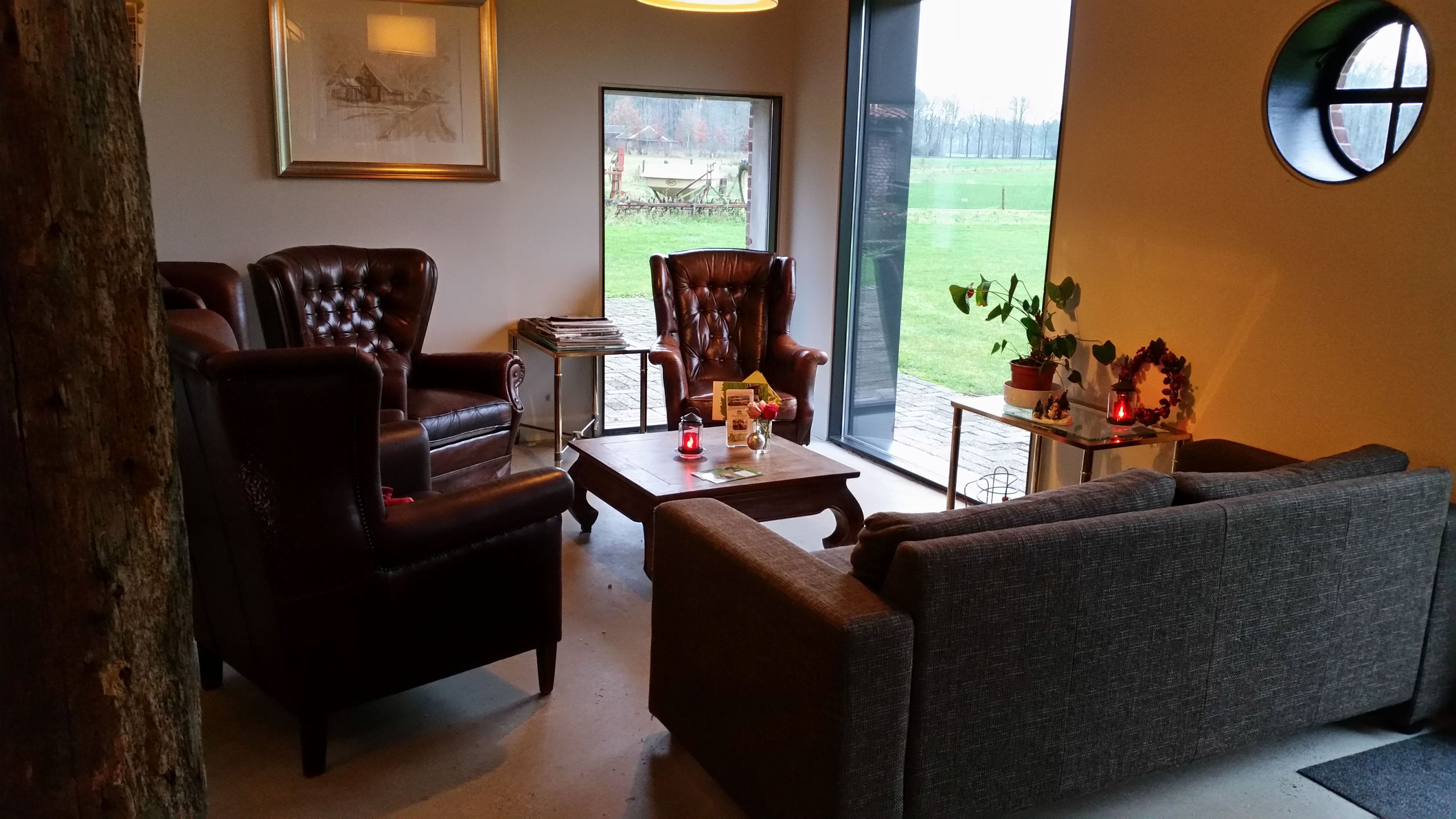 Ein gemütliches Wohnzimmer mit Ledersesseln, einem grauen Sofa, einem Couchtisch mit Buch und Kerze und Blick auf ein grünes Feld durch ein großes Fenster.
