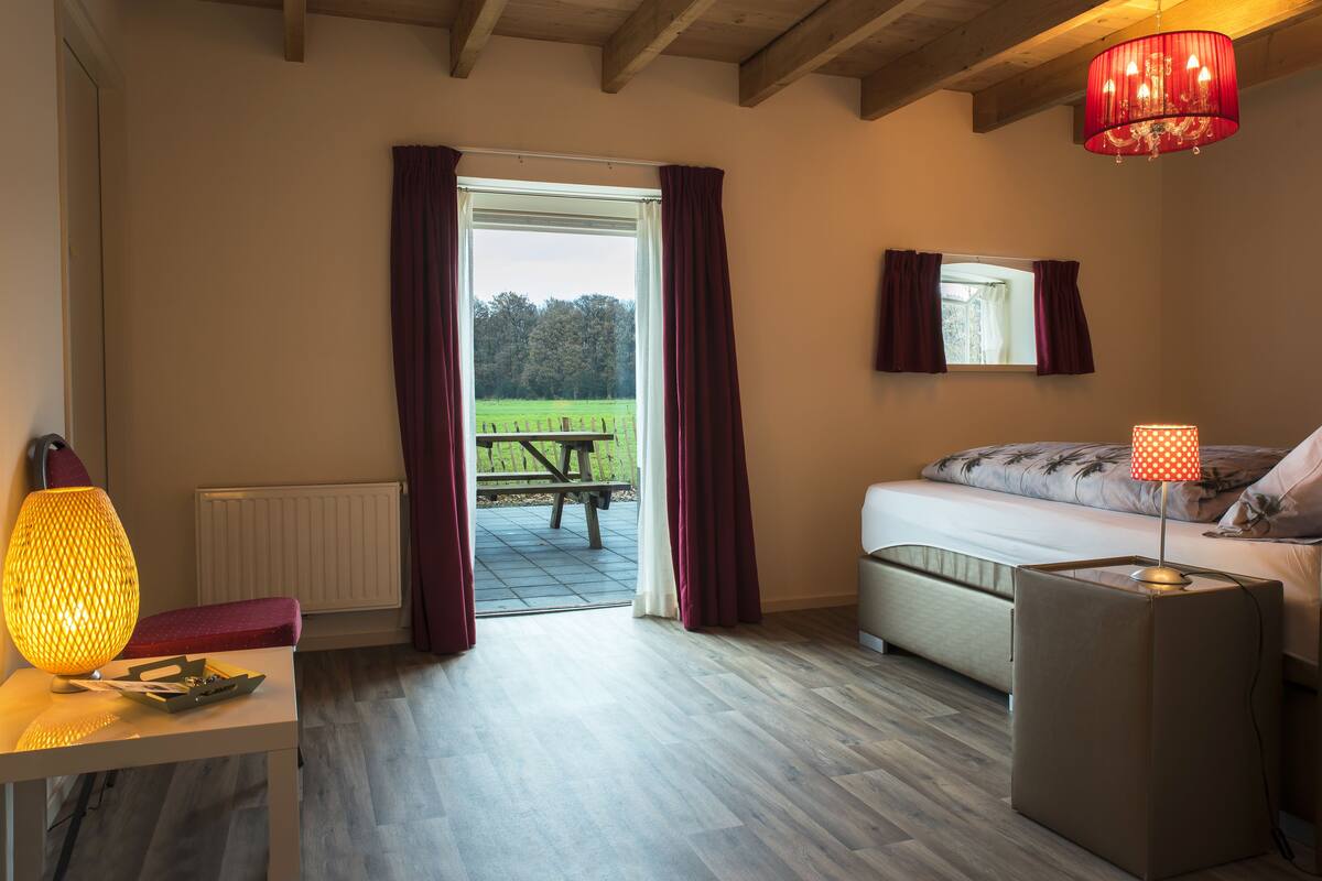 A room with a bed, a table with a lamp, a bench, and a view of a garden through a door.