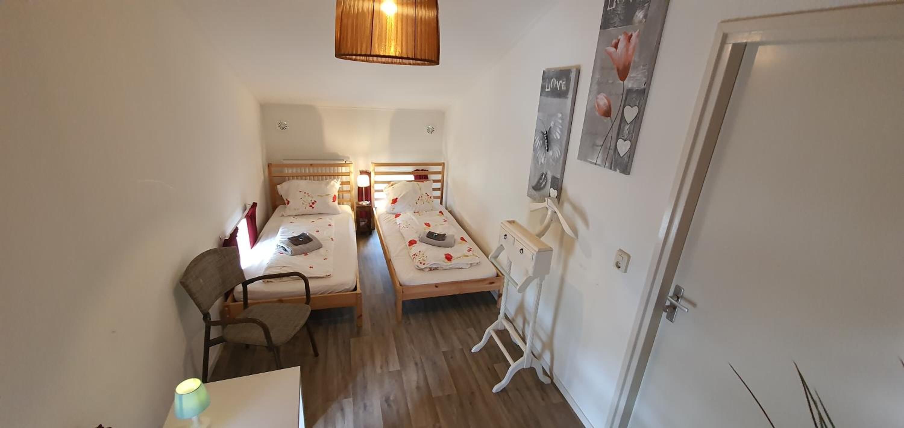 A small, compact room with two wooden beds, a desk, and a chair. There is a hanging light fixture above the beds and a door on the right side.