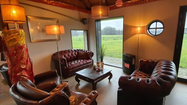 Una sala de estar con sofás de cuero, una mesa de centro y una chimenea. Amplias ventanas ofrecen vista a un campo verde.