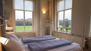 Habitación con cama individual, vista a un campo verde a través de amplias ventanas, una mesita de noche con lámpara y un radiador.