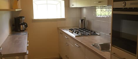 Een kleine keuken met houten kastjes, een gasfornuis, een koffiezetapparaat en een raam dat voor natuurlijk licht zorgt.