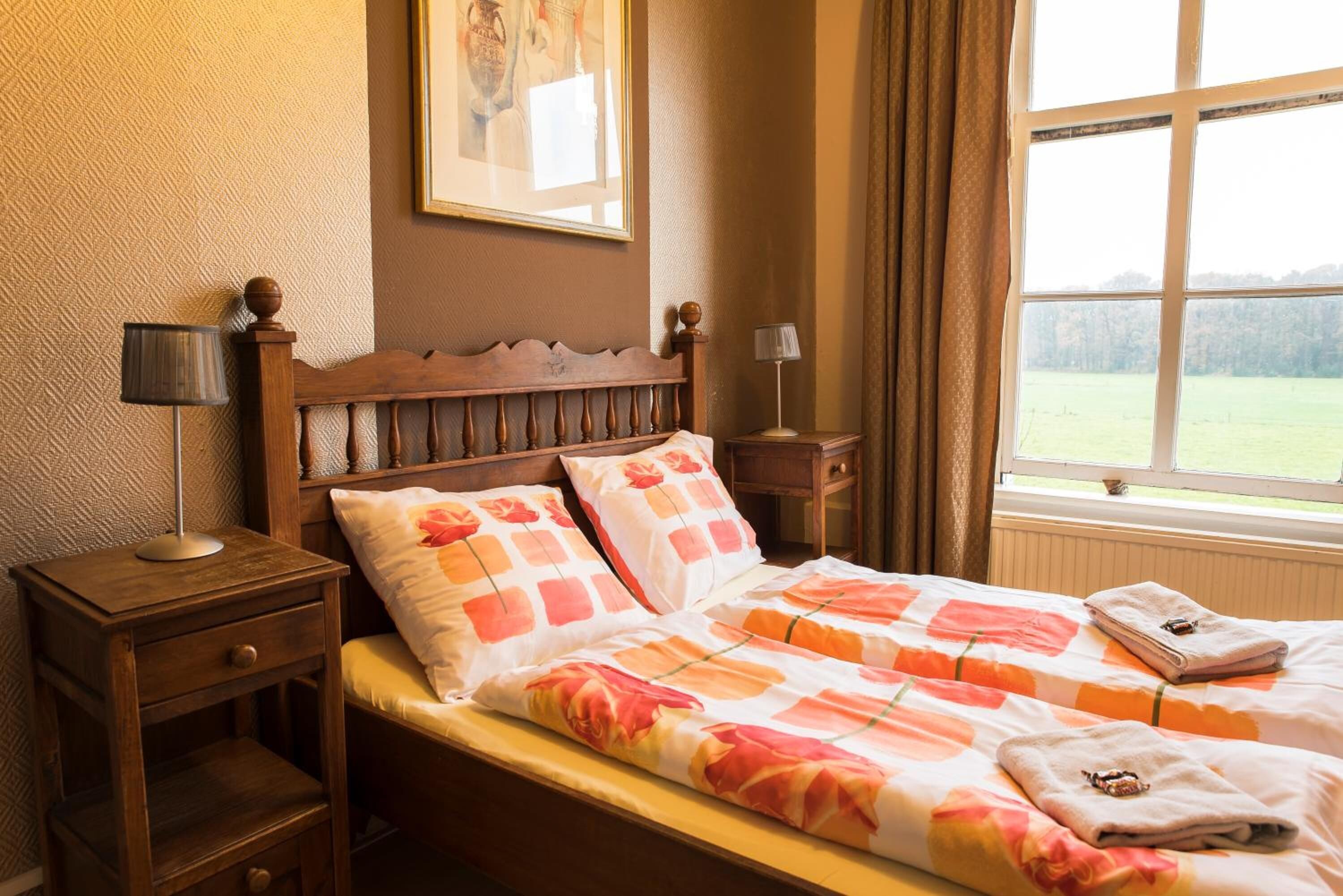 A bedroom with a wooden headboard, two bedside tables with lamps, a bed with floral bedding, and a window with a view of a green field.
