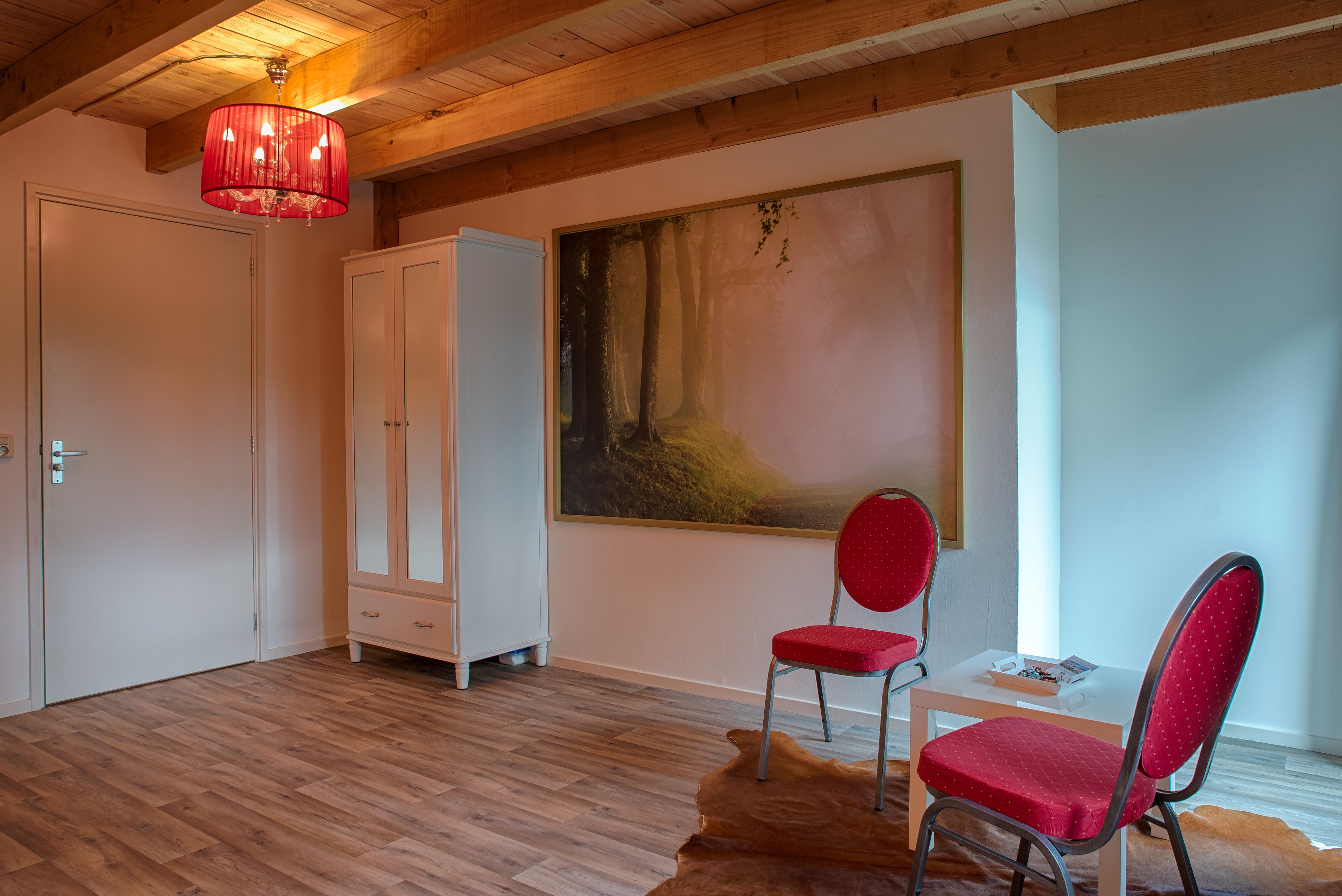 A room with wooden flooring, a red chair, a white cabinet, and a painting on the wall.