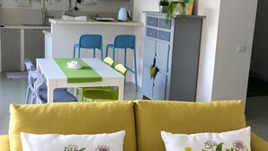 Een moderne woonkamer met een gele bank, groene en witte streep verlichtingsarmaturen, een eethoek met blauwe stoelen, en een keuken op de achtergrond.
