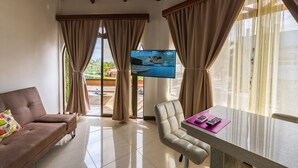 A hotel room with a sofa, a chair, a table with a remote control, and a TV displaying a landscape. - In the heart of the city (La Fortuna)