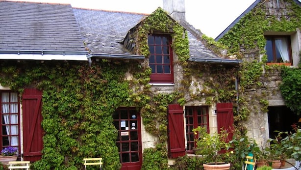 A stone building with ivy-covered walls, red shutters, and a tiled patio with outdoor furniture. - Chambres d'Hôtes La Poirière (Brissac Loire Aubance)