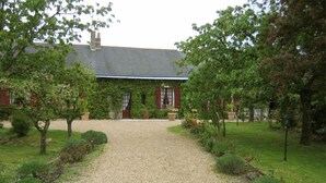 A building with a gravel driveway leading to a red brick entrance, surrounded by well-maintained greenery and trees. - Chambres d'Hôtes La Poirière (Brissac Loire Aubance)