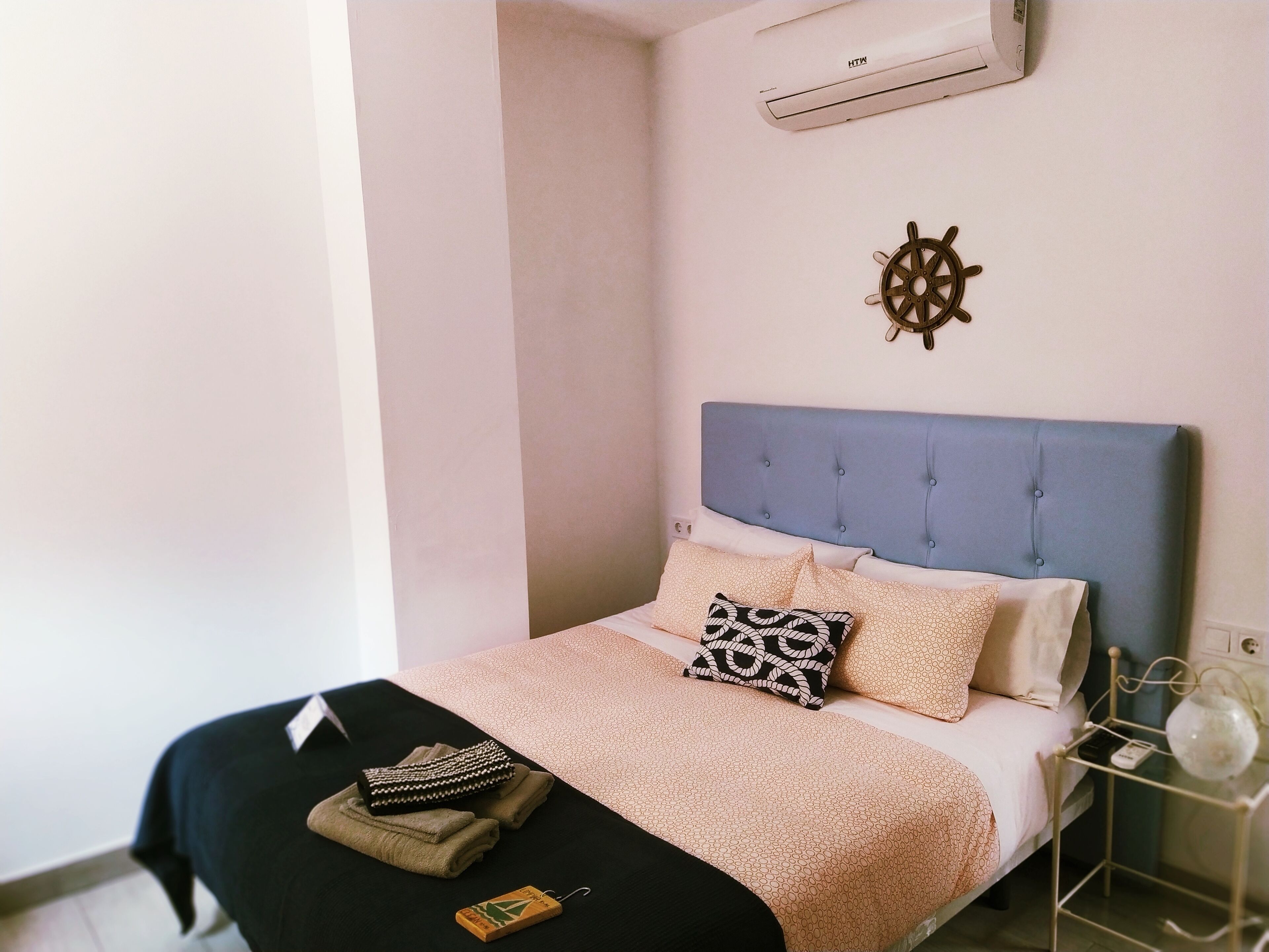 A neatly made bed with a patterned bedspread, a decorative ship's wheel, and a small bedside table with a glass vase.