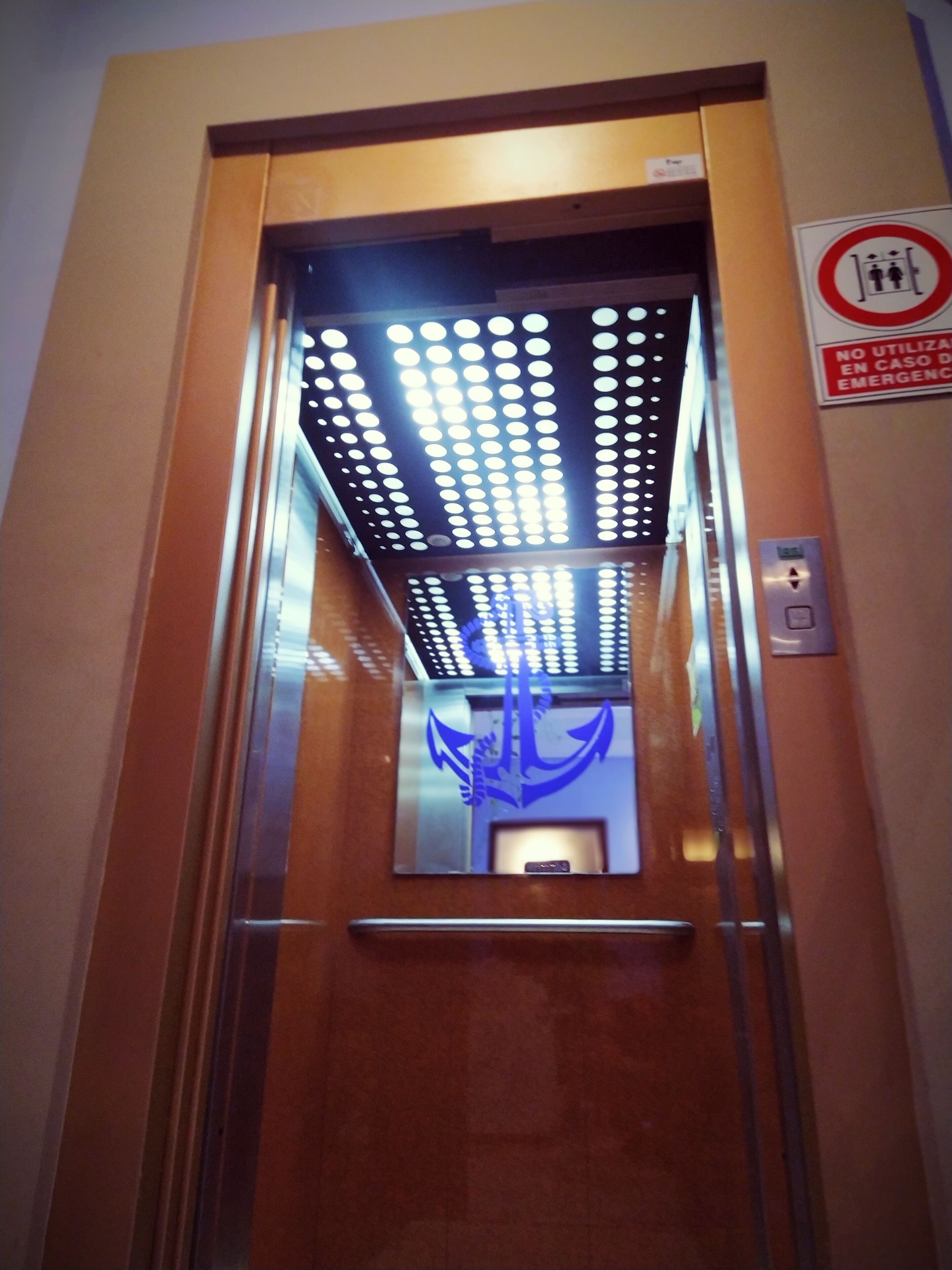 Interior de un ascensor con un símbolo de ancla azul en la pared, un cartel de prohibido fumar y un panel de botones.