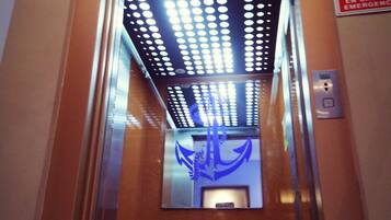 Interior de un ascensor con un símbolo de ancla azul en la pared, un cartel de prohibido fumar y un panel de botones.