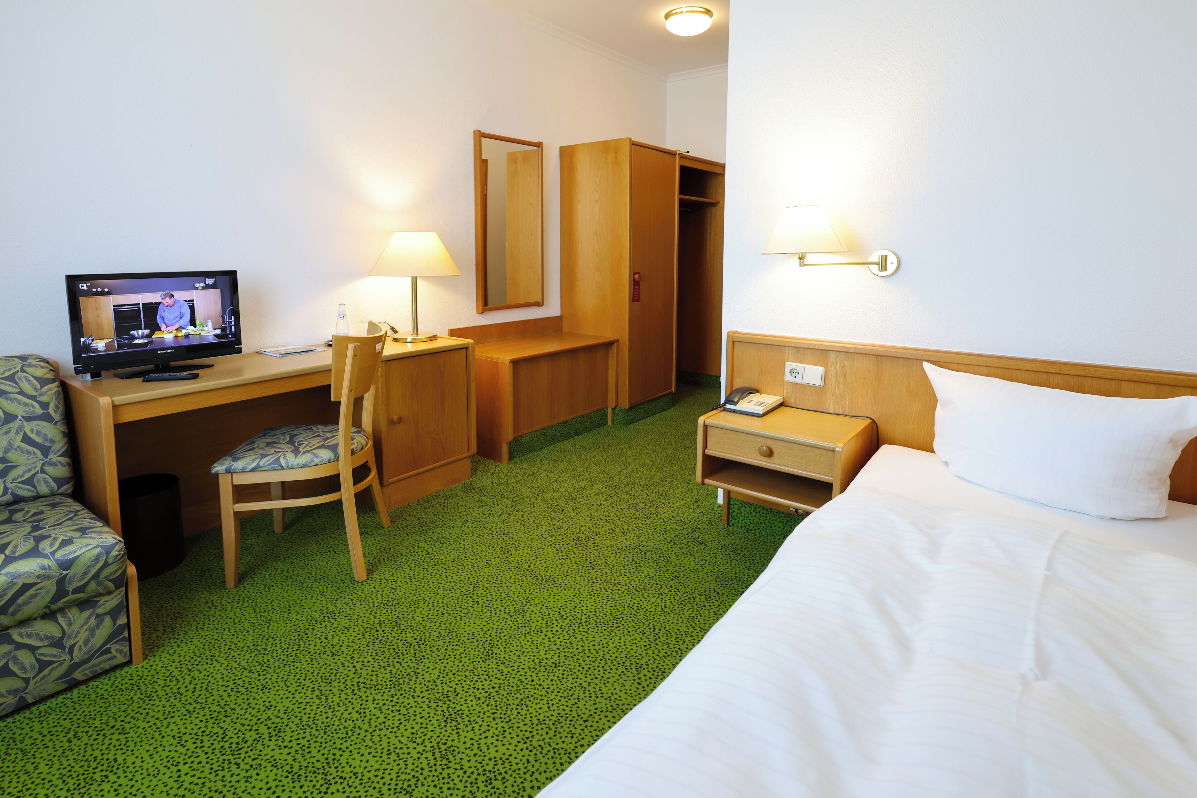 A hotel room with a bed, a desk with a computer, a chair, a wardrobe, and a bedside table with a lamp.