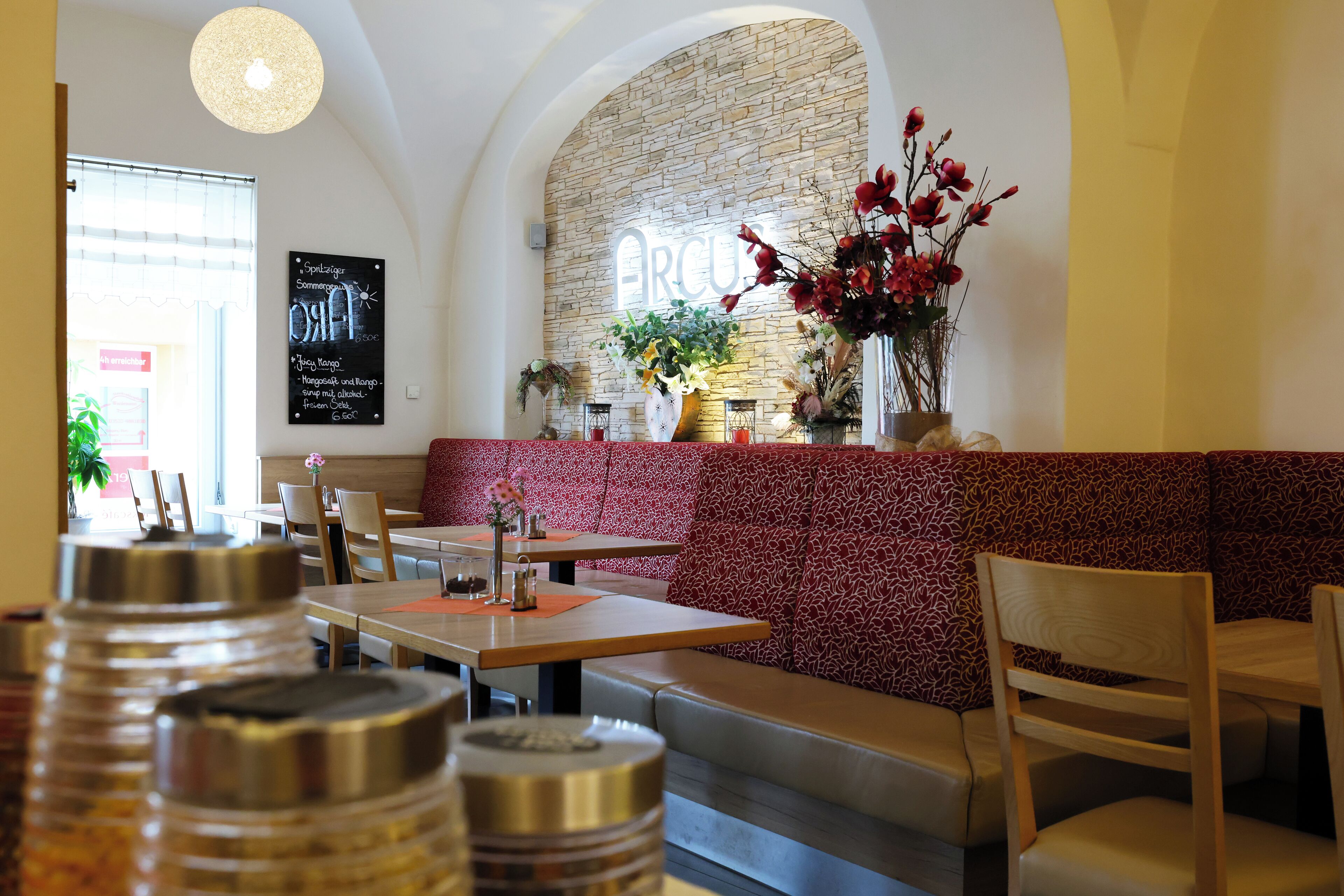 Interior de restaurante con pared de piedra, un banco de color rojo con estampado y un arreglo floral colgante.