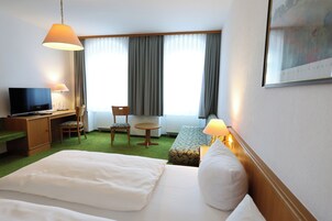 Quarto de hotel com cama, escrivaninha com televisão, cadeira, mesinha e janela com cortinas.