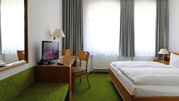 Quarto de hotel com uma cama, uma escrivaninha com cadeira, uma televisão e duas janelas com cortinas.