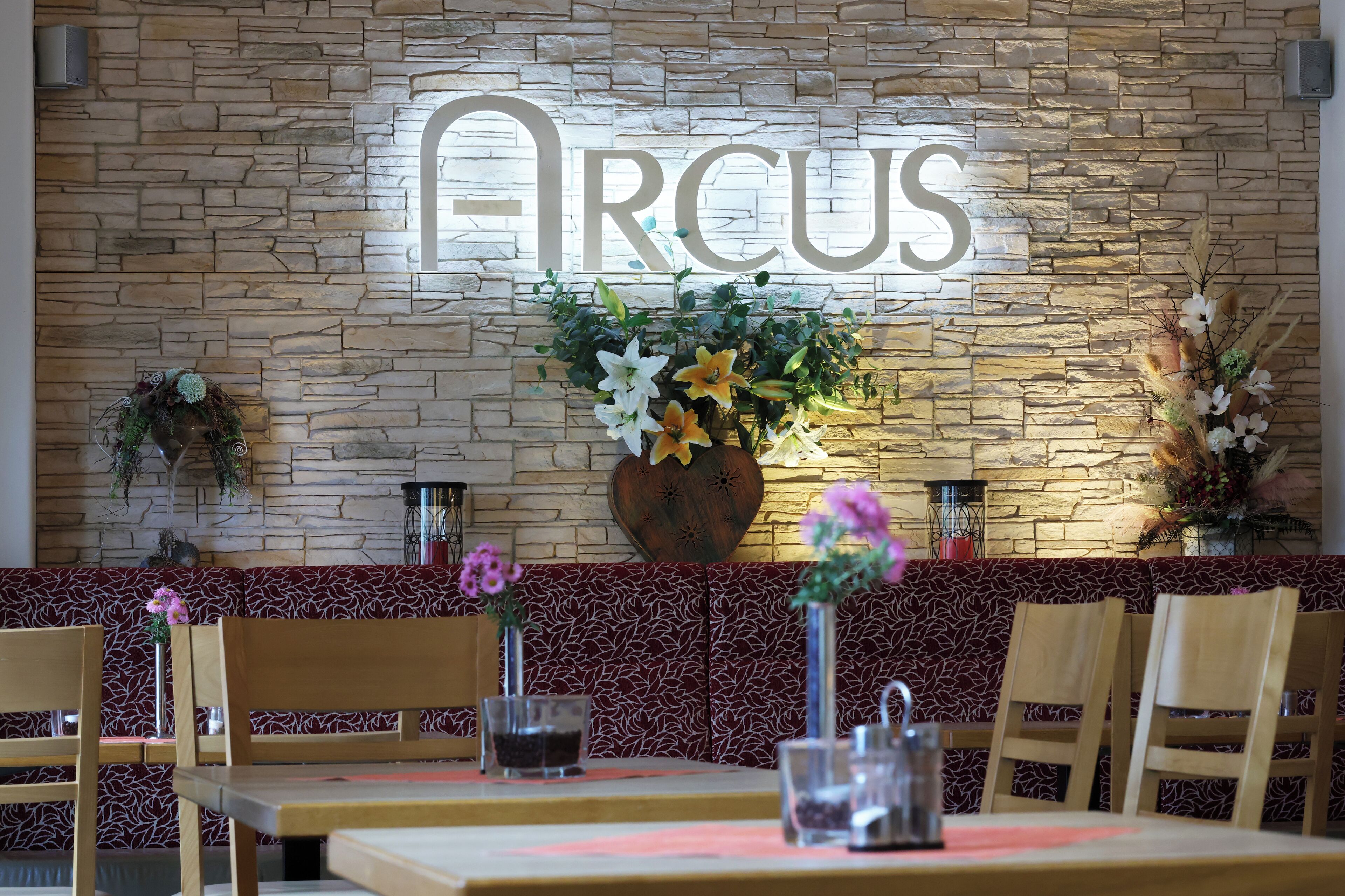 Interior de restaurante con pared de piedra, un letrero de neón que dice 'ARCUS' y adornos florales.