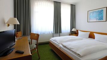 A hotel room with a bed, a desk with a TV, a chair, and a window with curtains.