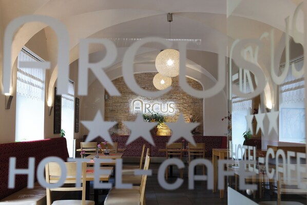A hotel cafe interior with a stone wall, wooden furniture, and a neon sign reading "ARCUS".