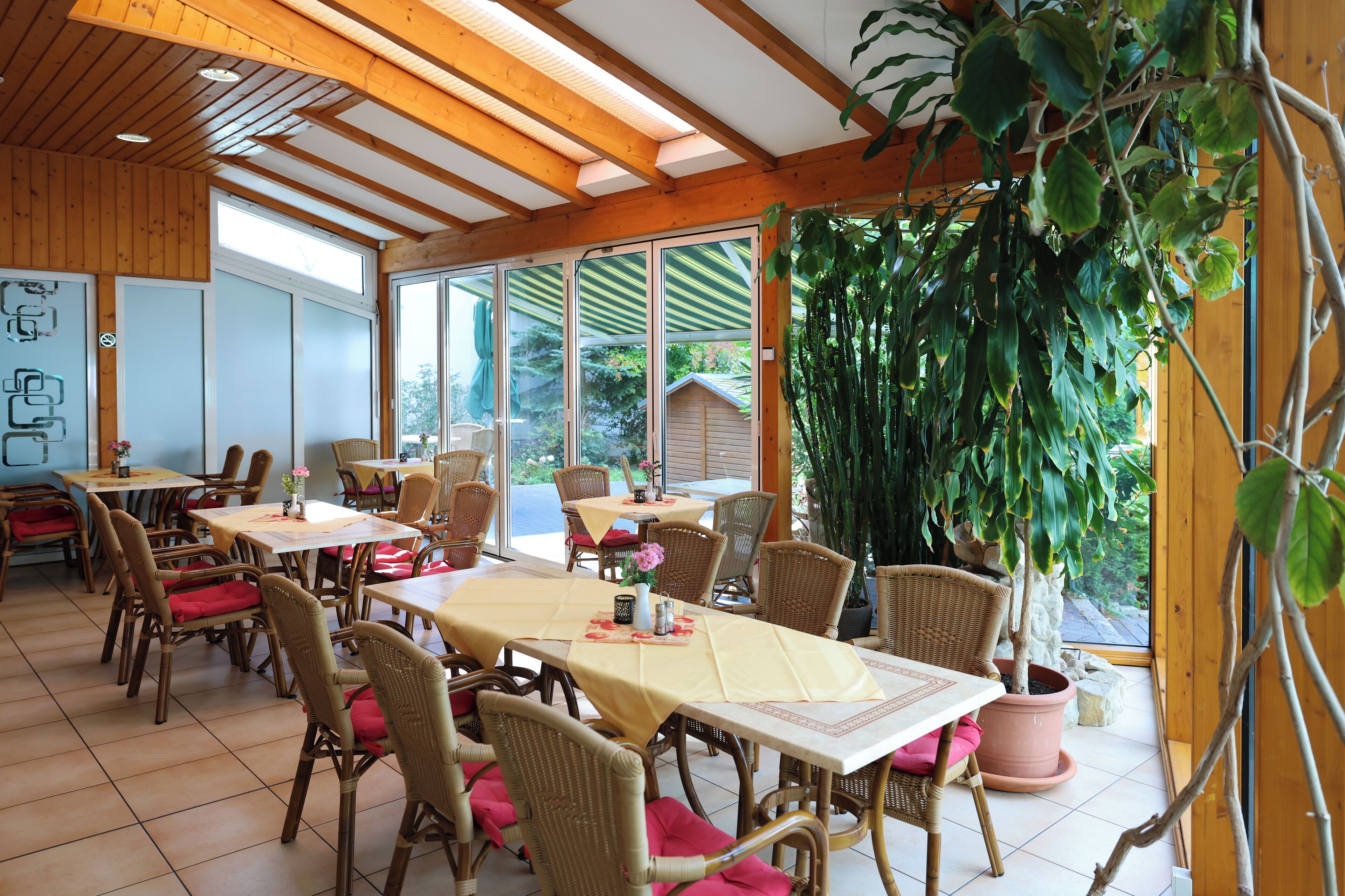 A dining area with wicker chairs and tables, large windows, and a potted plant.